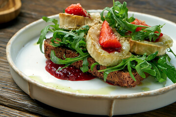 Appetizing toast with goat cheese, red jam, arugula and strawberries with rye bread on a plate on a wooden background