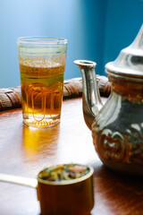 vertical image of near an antique metal teapot with a glass cup full of tea, and a spoon with dry grass on a wooden table.