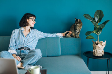 Beautiful brunette with a short haircut works in the home office on a blue background. Typing on a laptop, calling on the phone, transferring documents, making notes, drinking coffee from a mug.