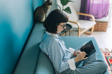 Beautiful brunette with a short haircut works in the home office on a blue background. Typing on a laptop, calling on the phone, transferring documents, making notes, drinking coffee from a mug.