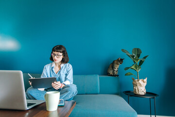 Beautiful brunette with a short haircut works in the home office on a blue background. Typing on a laptop, calling on the phone, transferring documents, making notes, drinking coffee from a mug.