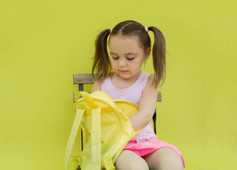 photo of a gorgeous, cute little girl in a skirt sitting isolated on a yellow background holding a backpack on her knees and looking into a yellow bag