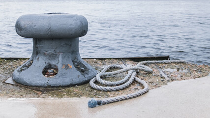 Fototapeta na wymiar Old mooring bollard at a dock with a rope lying next to it. Grey water in the background.