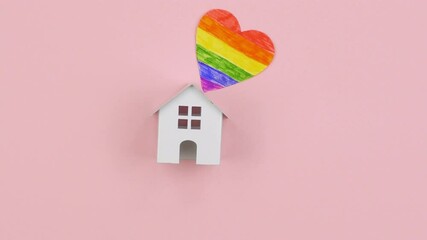 Female hand in an LGBT bracelet puts toy house, rainbow heart and ribbon on a pink background. Tolerance to homosexual and bisexual people community concept.  LGBT pride. - Powered by Adobe