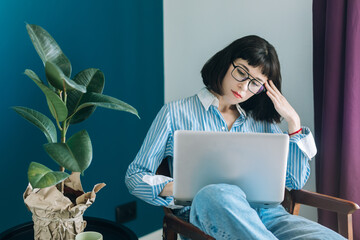 Beautiful brunette with a short haircut works in the home office on a blue background. Typing on a laptop, calling on the phone, transferring documents, making notes, drinking coffee from a mug.