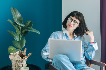 Beautiful brunette with a short haircut works in the home office on a blue background. Typing on a laptop, calling on the phone, transferring documents, making notes, drinking coffee from a mug.