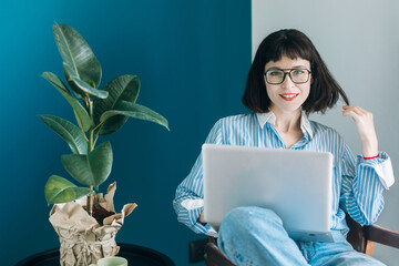 Beautiful brunette with a short haircut works in the home office on a blue background. Typing on a laptop, calling on the phone, transferring documents, making notes, drinking coffee from a mug.