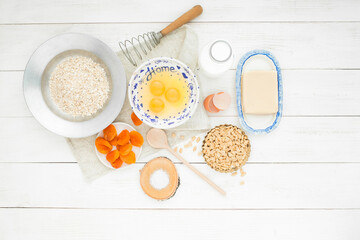 Raw eggs in a bowl, butter, milk bottle, peanuts, whisk, spoon, bowl of cereal, dried fruits on a light wooden table against a white background. The ingredients for the cookies.
