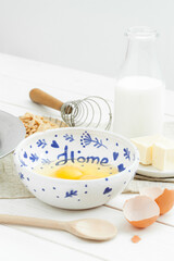 Raw broken eggs in a bowl, butter, milk in the bottle, peanuts, whip, spoon on a light wooden table against a white background. Ingredients for cookies.