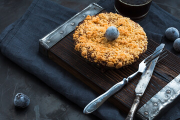 Cake and frozen plums on a wooden tray and a cup of coffee, knife and fork on a dark metallic background.