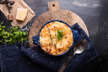Lasagna in baking dish on an old chopping board, basil, old spoon, blue napkin, parmesan, rosemary, wine opener on a dark metal table. View from above. Italian food.