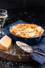 Lasagna in baking dish during eating carelessly spilled cheese, an old spoon, a glass of white wine, parmesan, rosemary on dark metal table. Close-up. Italian food.