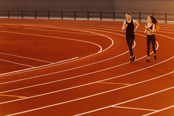 Young man and woman running on race track in modern stadium. 