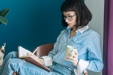 Beautiful brunette with a short haircut works in the home office on a blue background. Typing on a laptop, calling on the phone, transferring documents, making notes, drinking coffee from a mug.