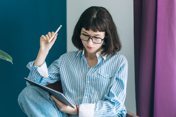 Beautiful brunette with a short haircut works in the home office on a blue background. Typing on a laptop, calling on the phone, transferring documents, making notes, drinking coffee from a mug.