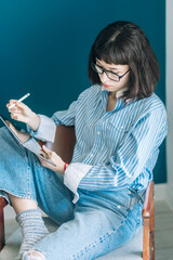 Beautiful brunette with a short haircut works in the home office on a blue background. Typing on a laptop, calling on the phone, transferring documents, making notes, drinking coffee from a mug.