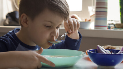 Cheerful boy eats soup with a spoon while sitting at the kitchen table. Hungry preschooler dines at home.