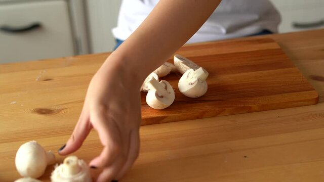Young woman cutting mushrooms on wooden board in kitchen. Female cutting white champignon with knife on chopping board on kitchen table.