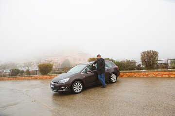 man stands near black car in rain in fog in city village Provence France. 