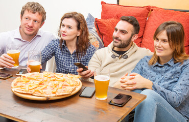 group of friends are drinking beer, eating pizza, talking and smiling while resting at home