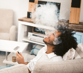 Shaggy Arab guy having fun smoking an electronic cigarette. Yiung man relaxing in a soft comfortable chair. Quarantine self-isolation entertainment concept. Toned image.
