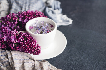 White cup with flowers on a saucer, a bouquet of pink lilac flowers and a blue drape striped scarf on a dark background, closeup, copy space