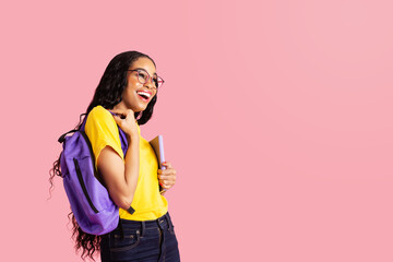 Portrait of a  laughing  young female student with book, backpack and glasses ready for school looking at  pink studio copy space
