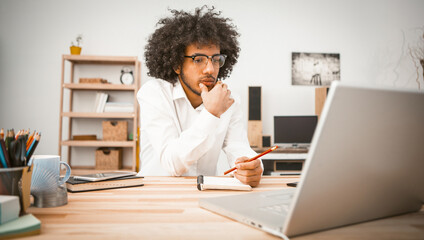 Pensive young man is engaged at home in front of computer. Arab student or businessman taking notes...