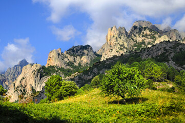 Fototapeta na wymiar Pins Laricio et Aiguilles de bavella en Corse, sur fond de ciel bleu nuageux et nature sauvage et abrupte des rochers formant une montagne découpée au profil accidenté et dentelé.