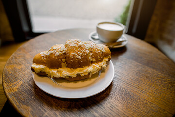 Fresh croissant with delicious cream on white plate and cup of coffee on the background, on round wooden table. Fresh aroma coffee with croissant on wooden table, front angle view