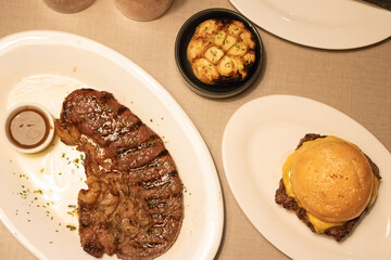 Medium grilled strip loin beef steak with pepper sauce, delicious cheese burger and garlic clove grilled in black bowl in warm light at steakhouse. Top view