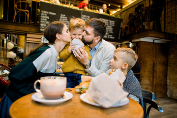 Happy lovely family, young pleasant parents and their two little sons, enjoying time together, sitting in cafe and having tasty lunch dinner with croissants. Mom and dad kissing their little son