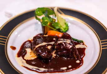 French main dish with stew of beef meat bourguignon simmered in red wine sauce with vegetables like carrots on a white and black dinner plate with golden rim.