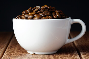 Coffee beans in a white cup over dark background and on a wooden surface