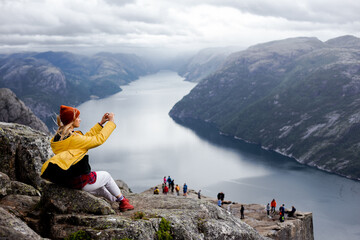 Dreamy blonde woman hiker in yellow raincoat sits on the top of the Preikestolen mountain...