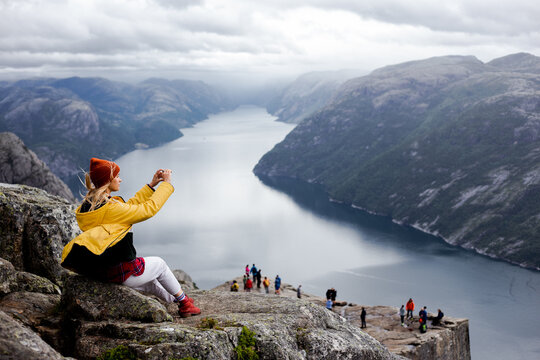 Dreamy blonde woman hiker in yellow raincoat sits on the top of the Preikestolen mountain (Preacher's Pulpit or Pulpit Rock) with a lot of tourists and shoots photos by phone on Lysefjord background