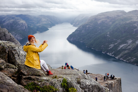 Smiling blonde woman hiker in yellow raincoat sits on the top of the Preikestolen mountain (Preacher's Pulpit or Pulpit Rock) with a lot of tourists and shoots photos by phone on Lysefjord background