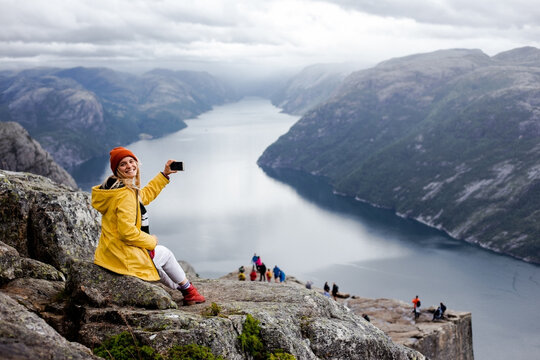 Smiling blonde woman hiker in yellow raincoat sits on the top of the Preikestolen mountain (Preacher's Pulpit or Pulpit Rock) with a lot of tourists and shoots photos by phone on Lysefjord background