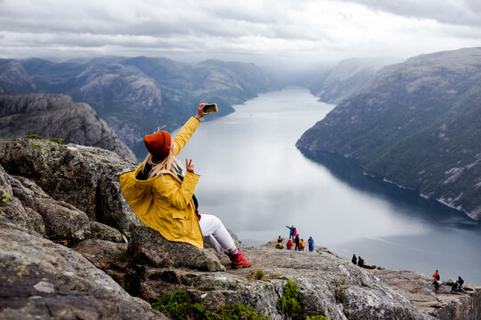 Happy blonde woman hiker in yellow raincoat sits on the top of the Preikestolen mountain (Preacher's Pulpit or Pulpit Rock) with a lot of tourists and shoots photos by phone on Lysefjord background