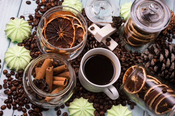 A white Cup of coffee on coffee beans, on a light blue wooden table. Wooden table. Fir cones on the table. Green marshmallows and white marshmallows. Dried citrus. Brown background.