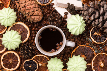 White Cup of coffee on coffee beans. light wooden table. Fir and cedar cones on the table. Green marshmallows and white marshmallows. Dried citrus. Brown background.