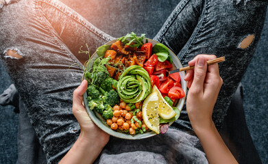 Woman in jeans holding Buddha bowl with salad, baked sweet potatoes, chickpeas, broccoli, greens,...