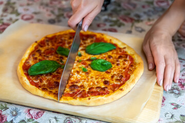 Close-up female hand cutting baked tasty hot homemade italian Margarita pizza on wooden cutboard and table with regular knife. Melted mozzarella cheese , tomato sausw and fresh green basil topping