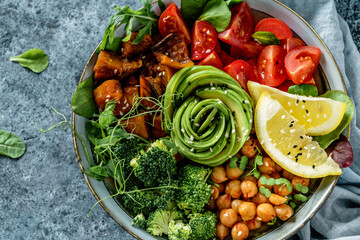 Buddha bowl salad with baked sweet potatoes, chickpeas, broccoli, tomatoes, greens, avocado, pea sprouts on light blue background with napkin. Healthy vegan food, clean eating, dieting, top view