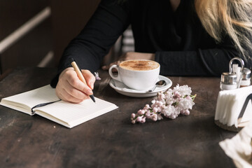 A girl in a coffee shop next to a cup of cappuccino writes in a notebook. Dark wooden table by the window and lilac