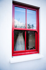 Beautiful white new wedding dress hanging behind the window, red painted wooden window frame, white wall, blue sky reflection in the window glass