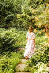 a girl in a long beautiful dress barefoot along the path goes into the forest. there are many flowering plants, shrubs, trees. There are stones near the gangway. Woman with her back to the photo