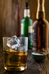 A glass of whiskey with ice, in the background are bottles on a wooden table of a bar counter, shallow depth of field, selective focus. The concept of alcoholic drinks in a roadside bar.