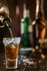 An alcoholic drink at the bar counter is poured into a tequila glass with salt at the edges, in the background are different bottles of alcohol, shallow depth of field, selective focus. The concept of