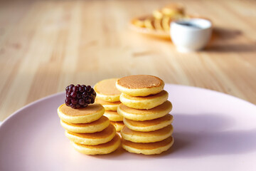 Trendy tiny pancakes cereal meal on pink plate. Baked pancakes for breakfast on wooden background. Fresh berries for decoration in background.